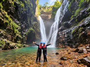 Duas mulheres sorridentes com roupas de neoprene, coletes salva-vidas e capacetes azuis, celebram diante da imponente Cachoeira do Dragão, cercada por paredões rochosos na Chapada dos Veadeiros, Goiás.
