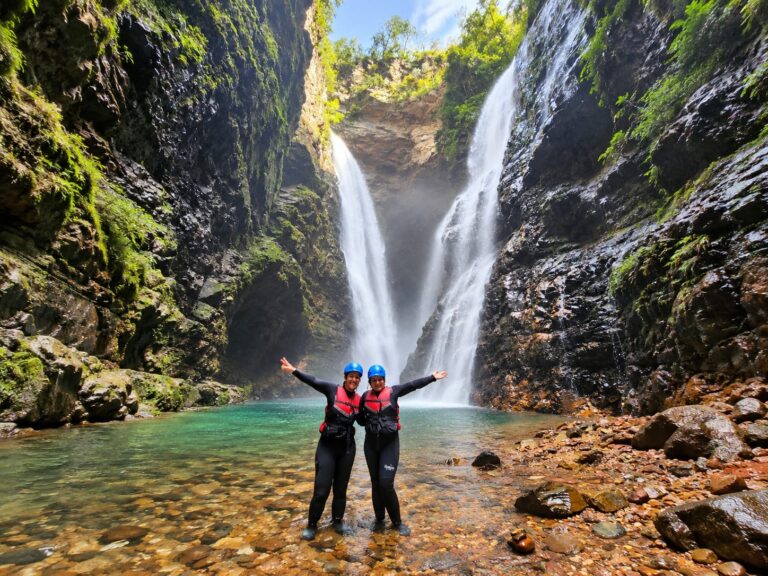 Duas mulheres sorridentes com roupas de neoprene, coletes salva-vidas e capacetes azuis, celebram diante da imponente Cachoeira do Dragão, cercada por paredões rochosos na Chapada dos Veadeiros, Goiás.