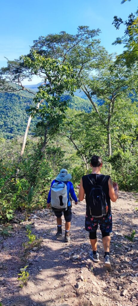 Dois visitantes com mochilas percorrem a trilha terrestre da Cachoeira do Dragão, uma descida íngreme cercada pela vegetação do cerrado, na Chapada dos Veadeiros